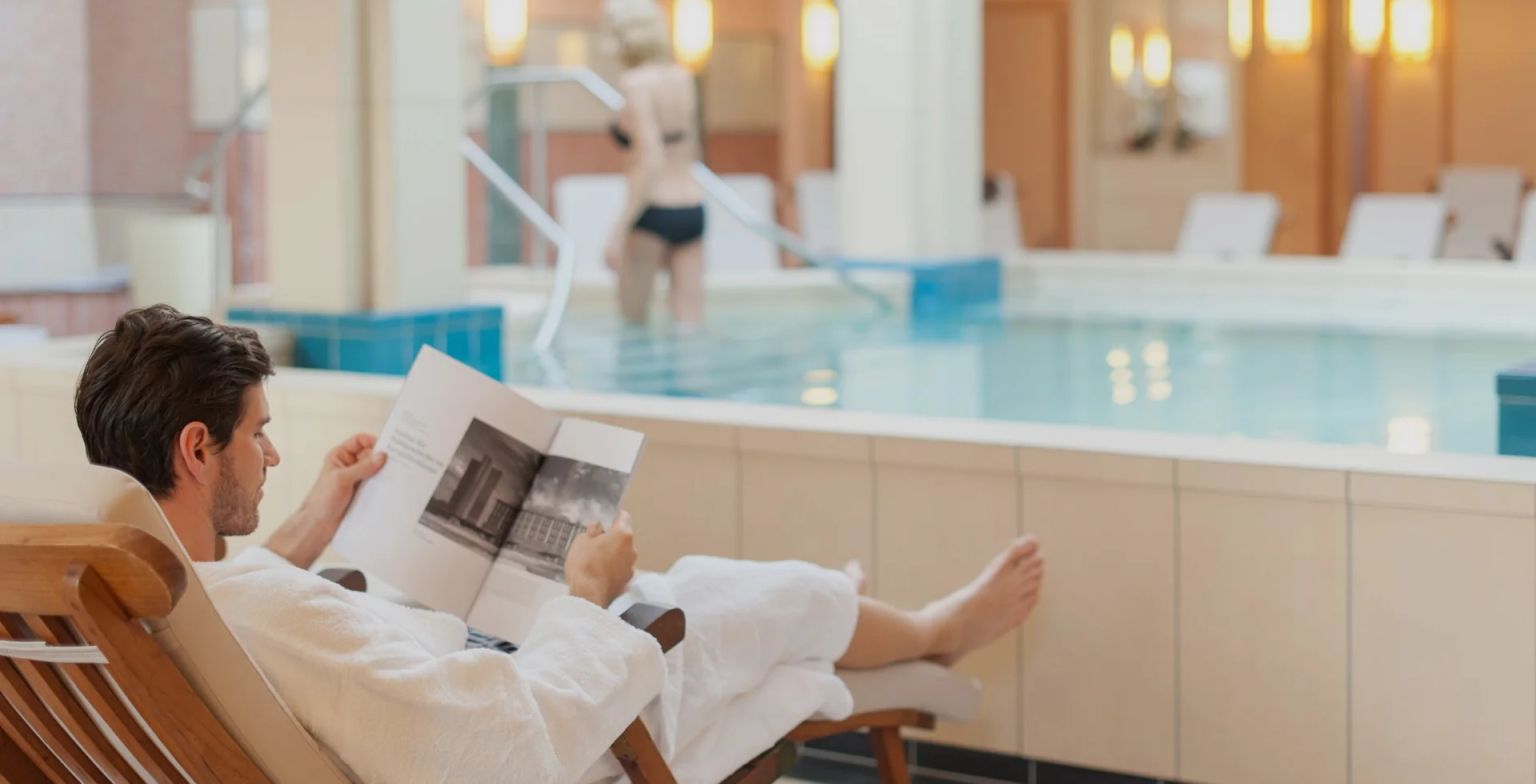 Kur-Royal Day Spa Bad Homburg Ein Mann liegt im Bademantel auf einem Liegestuhl vor einem Indoor-Pool und liest